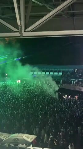 The video captures a vibrant outdoor concert in Sarajevo, Bosnia and Herzegovina at night. A massive crowd of people fills the space, with many illuminated by green and purple lights and lasers. The atmosphere is energetic, with a stage featuring a large screen displaying abstract visuals. Smoke machines are in use, creating a dramatic effect with the colored lights. The audience appears engaged and excited, with some holding up their phones, likely to record the event. The background shows tiered seating and the skeletal structure of the venue's roof, typical of an outdoor arena or stadium. There are visible signs with the word "rebel" written in a stylized font, suggesting a sponsorship or brand presence at the event. The overall mood is one of festivity and excitement, characteristic of a large-scale music performance.