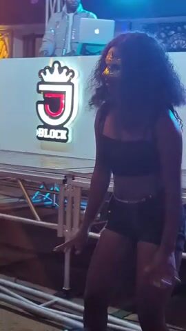 A young woman with dark, curly hair is dancing in front of a DJ booth. She is wearing a black crop top and black shorts. The DJ booth has a sign that says "J BLOCK" with a crown above the "J". There is a laptop and other DJ equipment on the booth. The background appears to be outdoors at night with lights visible.