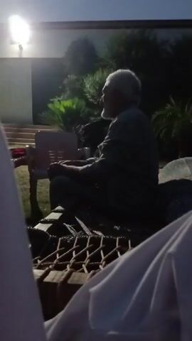 The video depicts an elderly man with white hair and a beard sitting on a woven bed outdoors at night. He is wearing a dark colored shirt and appears to be gesturing with his hands while speaking. Behind him is a building with a bright light shining on it and various plants. In the foreground, there are white garments that suggest the presence of other people. The scene is dimly lit, creating a silhouette effect for the man on the bed.