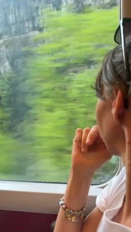 The video shows a woman riding on a train, looking out the window at the scenery. She is wearing a white t-shirt and a bracelet with charms. The train is moving, and the landscape outside is blurred. The scenery consists of green trees and a rocky cliff face. The sky is overcast. There are reflections on the train window.