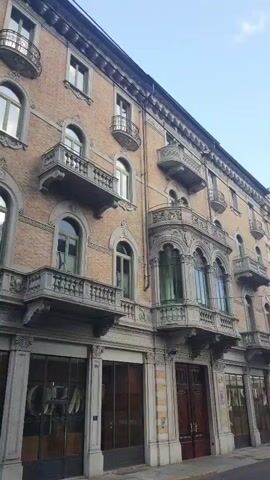The video shows a close-up view of a building facade with ornate architectural details. The building appears to be made of brick and stone, with multiple stories and balconies. The balconies have decorative railings and elaborate carvings. The windows are arched and some have green tinted glass. The ground floor has storefronts with large glass windows and wooden doors. The sky is visible at the top of the frame and appears to be a clear blue. There are no people or cars visible in the video. The camera is stationary and focuses on a specific section of the building.