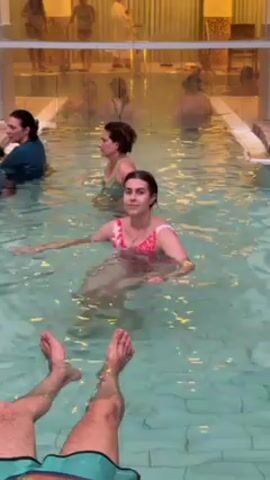 The video shows an indoor pool area with several people in the water. In the foreground, a pair of legs with green and blue patterned swim trunks are seen extended into the water. Three women are visible in the pool, with the woman nearest to the camera wearing a pink floral swimsuit. She is floating on her back and looking towards the camera with a smile. Behind her, two other women are also in the water. The pool is surrounded by a tiled deck, and a glass partition with more people on the other side is in the background. The overall lighting is bright and the water appears clean.