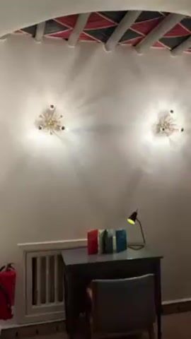 The video shows a slow pan of a hotel room interior. It begins with a view of a small desk with books and a lamp, flanked by two light fixtures on the wall.  The camera then moves to the right, revealing a double door closet. The pan continues to show a bed with white linens and a rug on the wall. An air conditioning unit is mounted high on the wall above the bed.  The ceiling has a dark wooden beam structure with colored inlays between the beams. The video ends with a partial view of an adjoining room with a chair, fireplace, and black and white art on the wall.