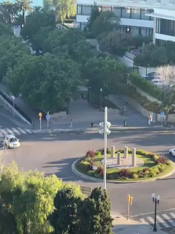 The video presents an elevated view of an urban landscape in Tarragona, Spain, during what appears to be a clear, sunny day. The scene centers around a bustling roundabout with a landscaped central garden, featuring decorative pillars and shrubbery. Several white and light-colored cars are observed navigating the streets and the roundabout, while pedestrians walk along the sidewalks and cross roads.

Lush green trees densely line the avenues, creating a verdant canopy. Multi-story white buildings, indicative of modern coastal architecture, frame the background. As the camera slowly pans to the right, the expansive blue sea becomes prominent in the distance, emphasizing the city's coastal setting. A flag, likely the Catalan Senyera with its distinctive red and yellow stripes, is visible on a pole to the left, confirming the regional context. The overall atmosphere is one of calm activity in a well-maintained and scenic environment. No discernible text is visible on signs or buildings.