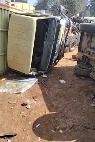 The video shows a scene of two overturned vehicles. The first vehicle is a light-colored truck with the name "BAMRA CEMENT" printed on it. It is lying on its side, with its cab open. The second vehicle, a car, is flipped onto its roof, exposing the undercarriage. A tow truck is visible in the background. There are people present, including one man in a black shirt who passes in front of the camera and another person wearing a blue and white vest standing beside a wooden fence. The ground is dirt, with scattered debris and small rocks. The video seems to be taken at a salvage yard or accident recovery site.