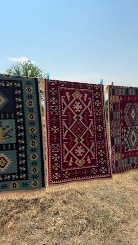 The video shows a row of intricately patterned rugs hanging on a clothesline outdoors. The rugs are suspended by clothespins against a clear blue sky. They are made of woven fabric, with geometric designs and fringe along the bottom edges. The rugs appear to be drying or airing out in the sun. In the background, there is dry grass and foliage, suggesting a rural or outdoor setting. A flagpole with a flag, possibly the Armenian flag, is visible in the distance. The scene is bathed in bright sunlight, casting shadows on the ground. The camera pans across the rugs, showcasing their detailed craftsmanship and vibrant colors.