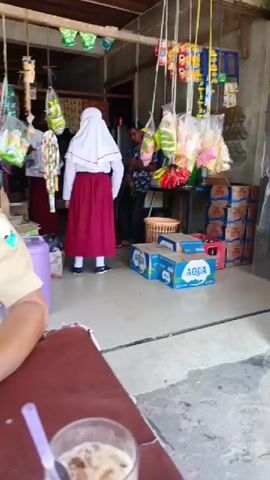 A static, eye-level shot shows the interior of a small shop. In the foreground, a glass of iced coffee with a spoon inside sits on a brown surface, presumably a table. Beyond this, the shop is open to the outside, with a variety of items hanging from the ceiling, including snacks and decorations. Several cases of bottled water, labeled "AQUA", are stacked on the floor. A woman in a white hijab and red school uniform stands in the center of the frame, facing towards the shopkeeper, who is partially obscured behind a table. Another girl in a school uniform is partially visible to the left. The shopkeeper, a man with dark hair wearing a patterned shirt, appears to be assisting the girls. A wooden chair is positioned on the right side of the frame. The floor is concrete. The overall atmosphere is casual and everyday.