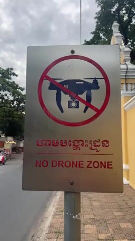 The video begins with a close-up of a sign indicating a "NO DRONE ZONE" in both Khmer and English. The sign features a red circle with a diagonal line through it, superimposed over a silhouette of a drone. The background shows a street scene with a cloudy sky. The camera then pans to the left, revealing a busy intersection with several people on motorcycles and scooters. Various small shops and food stalls line the street, with trees providing shade. A few vehicles, including tuk-tuks, are also present. People are seen interacting, with some riding on motorcycles, others sitting at stalls, and a few walking. The overall environment appears to be an urban or semi-urban area, possibly in Southeast Asia given the script on the sign and the presence of tuk-tuks. The footage captures everyday activity in a public space where drone operation is prohibited.