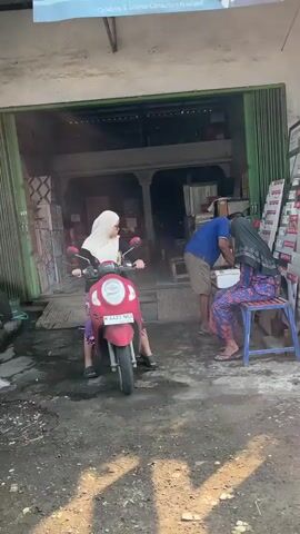 A woman wearing a white hijab, sunglasses, and a patterned skirt is seated on a pink scooter. The scooter is positioned on a paved area in front of a building. To her right, another woman in a blue and purple patterned outfit and a dark hijab sits on a blue stool. A third person, wearing a blue shirt and dark pants, is standing and appears to be interacting with the seated woman on the stool. The background shows the interior of a store with stacks of tiles and other merchandise. The storefront has a large green corrugated metal door that is partially open, and a green metal frame above the entrance. The ground is wet and appears to be a mix of dirt and pavement. The overall environment suggests an outdoor commercial setting.
