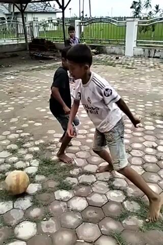The video shows a group of barefoot children playing soccer in an outdoor area. The ground is covered in hexagonal paving stones with patches of moss or grass growing in the cracks. There are three children visible who are actively involved in the game. One is wearing a white shirt and denim shorts, focusing on dribbling the soccer ball. Another is wearing a black shirt and shorts, running to keep up with the play. A third child is wearing a plaid shirt and red pants, walking into the frame. A building is visible in the background, with a corrugated metal roof and a wooden frame. A fence runs along the perimeter of the yard. The light is bright, suggesting it is daytime. The sound includes children speaking.