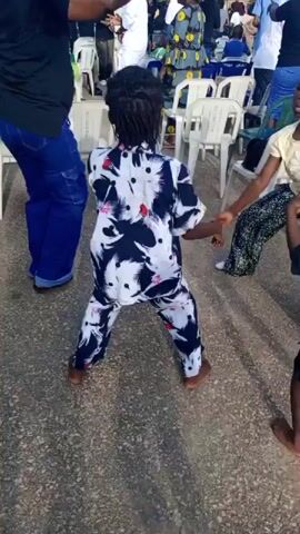 The video opens with a full shot of a child dancing with their back to the camera. The child is wearing a black, white, and pink patterned outfit. Their hair is in braids and they appear to be barefoot. They are dancing with their arms raised, and their body is in a bent-knee stance. The environment is outdoors with a paved surface. White chairs are visible in the background, arranged in rows, and several adults and children are present. The child continues to dance, turning slightly to the left and right. The camera then pans and zooms to the right, revealing more of the scene. Several other children and adults are visible. One child, wearing an orange shirt and patterned pants, is also dancing and appears to be interacting with the first child. Another child, wearing a denim jacket and jeans, is dancing in the center of the frame. Two girls are seen dancing on the left side of the frame, one wearing a yellow shirt and the other wearing a green shirt. The overall atmosphere appears to be a festive gathering or celebration.