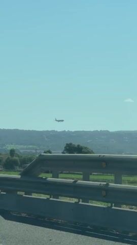 The video shows a bright, clear sky with an airplane flying in the distance. Below the airplane, there is a landscape with rolling hills covered in trees and vegetation. In the foreground, a highway barrier is visible, suggesting the video was filmed from a moving vehicle. The lighting indicates it is daytime.