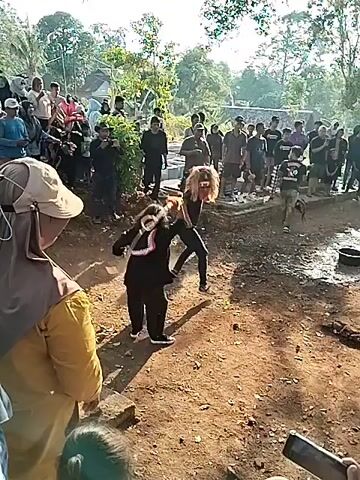 A crowd of people stands gathered outdoors, observing two individuals engaged in what appears to be a performance or a spirited competition. The ground is dirt and uneven. On the left, a person in black clothing, possibly a traditional outfit, holds a staff-like object with a round, orange object at the end. They are in a dynamic pose, mid-motion, with dust or debris kicked up around them, suggesting forceful movement. Facing them, on the right, is another individual wearing black pants and a dark t-shirt, with blonde hair. This person is also in an athletic stance, leaning forward with their arms raised, seemingly reacting to the action from the other individual. The crowd is attentive, with many people looking towards the central action. The background shows vegetation and trees, indicating a rural or natural setting, possibly a village or a park. The lighting suggests it is daytime, with sunlight casting shadows.