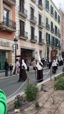 The video shows a procession on a city street. People wearing hooded robes, some white and some brown, walk slowly in formation, some carrying a cross or a banner.  People are standing on the sidewalk watching. Buildings line the street on both sides.