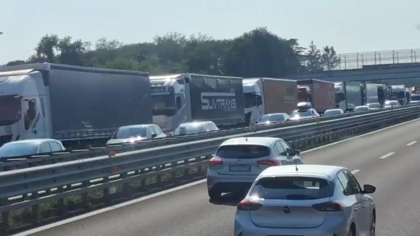 The video shows a traffic jam on a highway. Numerous trucks and cars are stopped on the road. The vehicles are lined up in multiple lanes, indicating significant congestion. A digital sign above the highway displays text in Italian, which appears to be related to traffic conditions, specifically "RALLENTAMENTO PER TRAFFICO INTENSO" (Slowdown due to heavy traffic). The environment suggests a sunny day with trees visible in the background. The perspective is from a vehicle moving slowly within the traffic jam.