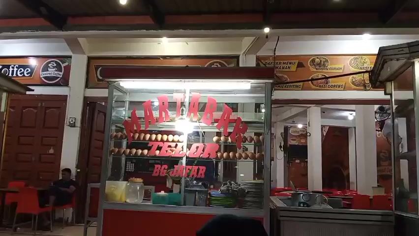 The video shows a street food stall at night. The stall has a sign that says "MARTABAK TELOR BG JAFAR". There are many eggs displayed in the stall. To the right of the martabak stall, there is another stall selling "MIE AYAM". There are customers sitting at tables in front of the stalls. The area is lit by overhead lights.