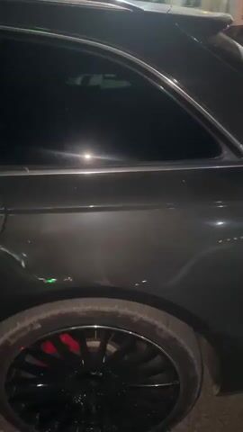 The video shows a dark-colored Audi A6 Avant parked at night. The camera is positioned low and to the side of the car, focusing on its rear quarter panel, taillight, and license plate. The license plate reads "MN EC 87 D". The car's paint is dark gray and appears to be slightly dusty. The taillight is illuminated with a red glow. The surrounding environment is dimly lit, with hints of streetlights and possibly building lights in the background. There are trees and a paved surface visible. The car is stationary throughout the short clip.