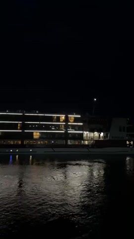 A night-time shot captures a cruise ship docked on the water. The ship is illuminated with rows of bright lights along its decks. The water below reflects the lights, creating a shimmering effect. The sky is dark, suggesting it is late at night.
