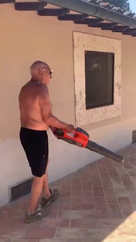 A shirtless, older man with a shaved head, wearing black shorts and grey sneakers, is using a red and black Husqvarna leaf blower. He is standing on a patterned terracotta tiled patio, facing away from the camera. The man is blowing air towards the wall of a building, which is painted a light beige color. A stone-framed window is visible on the building's wall. To the right of the building, there are raised wooden planters filled with greenery, and beyond that, trees and foliage are visible. The sun appears to be shining brightly, casting shadows on the patio, suggesting it is daytime. The scene takes place outdoors, likely in a residential or garden setting. The man's posture and the way he holds the leaf blower suggest he is actively engaged in clearing debris. There is no explicit emotion conveyed, but the activity suggests a task being performed. The visible text on the leaf blower is "Husqvarna". The location context provided is Castiglione d'Orcia, Italy, which suggests a Mediterranean climate and architectural style.