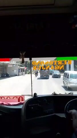 The video shows a view from the inside of a bus, looking out the windshield. The bus is driving on a paved road. In front of the bus are several vehicles, including a truck, a car, and a motorcycle. The landscape outside appears to be rural or suburban, with trees and buildings visible on the sides of the road. There is a sign on the windshield of the bus that is partially obscured, but some of the lettering is visible. The driver's hands are visible on the steering wheel. The video is shaky, suggesting that the bus is in motion.