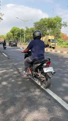 A motorcyclist wearing a dark blue shirt and a dark helmet rides on a paved road. The road has a white line marking the lane and a curb on the right side. Trees line the right side of the road, and a yellow truck is parked there. The sky is blue with some clouds. The footage is shaky, suggesting it was filmed from another moving vehicle.