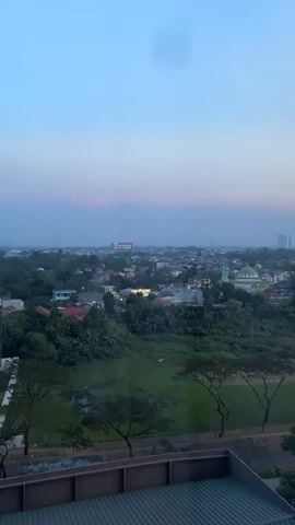 The video pans across a cityscape at dusk. The sky is a pale blue, transitioning to a soft pinkish hue near the horizon. In the foreground, the top of a building with a metallic, corrugated roof is visible. Beyond that, a patch of grass and some trees line a road. Further into the distance, a sprawling urban landscape unfolds, dotted with houses, trees, and a prominent green-domed building, possibly a mosque. The overall atmosphere is serene, suggesting the end of the day. No people or distinct actions are captured.