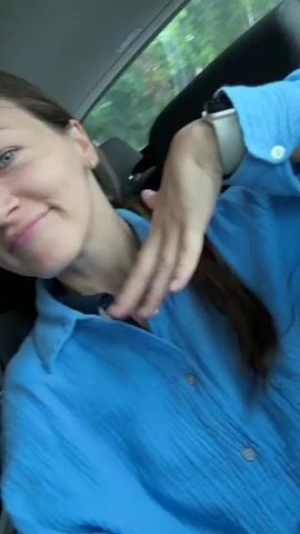 A young woman with long dark hair is seated inside a car, looking directly at the camera with a slight smile. She is wearing a light blue, collared, button-down shirt over a darker undershirt. A white smartwatch is visible on her right wrist. Throughout the video, she makes a casual, somewhat dismissive gesture with her right hand, moving it from near her chest downward and outward. Behind her, through the car window, blurred green foliage and what appears to be a blue circular road sign are visible. The car interior shows black upholstery.
