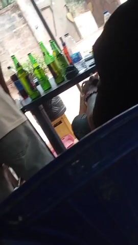 From a low-angle perspective, the video shows a man standing near a table with bottles of beer and soda. The table is black with metal legs, and there are several green beer bottles, a bottle of Pepsi, and other bottles on top. The man is wearing a light-colored shirt and grey pants. Another person is seated at the table, visible from the side. The setting appears to be outdoors, possibly a casual gathering or a bar.