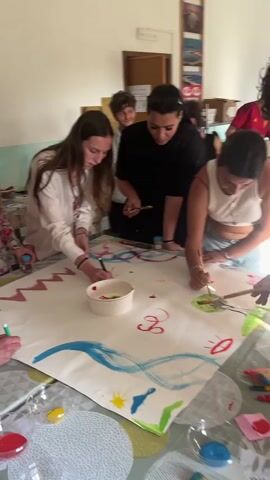 A group of eight young adults gather around a large white paper laid out on a table, creating a collaborative painting. Several people are actively painting on the paper, using various colors and brushes, while others observe. The table is covered with a patterned tablecloth and holds small containers of paint, paintbrushes, and markers. The room is well-lit, with a door visible in the background and pictures hanging on the wall. The overall atmosphere appears relaxed and creative.