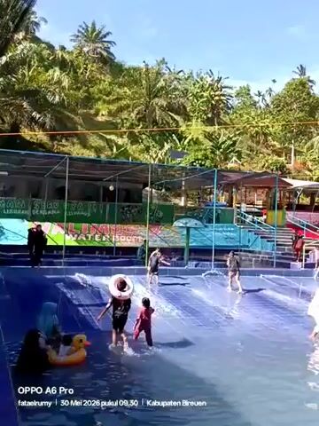 Here is a description of the video:

The video shows a shallow water park with multiple people, including children, enjoying the water. Some are walking, some are playing, and some are being helped by adults. The water park has a small water slide and an artificial mushroom fountain. The background features lush green trees under a clear blue sky.  Text at the bottom of the screen reads "OPPO A6 Pro fataelrumy 30 Mei 2026 pukul 09.30 Kabupaten Bireuen".