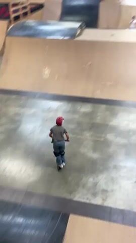 A person in a red helmet, gray t-shirt, and knee pads rides a scooter up a ramp in a skatepark. The person is in mid-air, performing a flip or trick. Other ramps and structures made of wood and black material are visible in the background, along with another person standing in the distance. The floor is concrete.