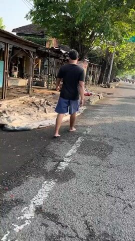 A person wearing a black t-shirt and blue shorts walks barefoot on a paved road. The person is walking away from the camera, towards the right side of the frame. The road is asphalt and has some cracks and white markings. On the left side of the road, there are several stalls with tarps spread on the ground. There are trees and buildings in the background. The lighting suggests it is daytime, possibly morning or late afternoon, with sunlight casting shadows. There are also some vehicles visible in the distance on the road.