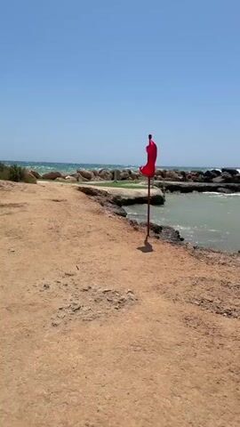 A red flag on a pole flutters in the wind near a rocky coastline on a sunny day. The flag is positioned on a sandy path leading towards the water, which is choppy with visible waves. In the background, there are more rocks and a faint silhouette of buildings against the clear blue sky. The camera is stationary, capturing the flag and the coastal scenery.