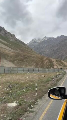 This is a first-person perspective video taken from inside a moving vehicle. The camera is pointed out the side window towards the mountain landscape. On the left side of the frame, a steep, barren mountain slope dominates the background, with patches of green vegetation near the base. A gray metal fence runs along the side of the road, separating the road from the mountain. The road itself is asphalt, with a yellow line marking the edge. In the foreground, a portion of the car's side mirror is visible, reflecting the interior of the car and the driver. The sky is overcast with heavy gray clouds. The video appears to be shot on a cloudy day, possibly in a mountainous region during daytime.