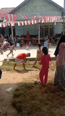 The video shows an outdoor scene in what appears to be a village gathering. A group of children are engaged in a game, possibly a race or competition, in the foreground. One boy in a red shirt and patterned shorts is bent over, as if about to throw or retrieve something small. Another child in a pink outfit stands nearby, watching. Several other children are visible in the background, some participating and others observing.

A house or building serves as the backdrop, decorated with red and white flags, suggesting a celebration or national holiday. Several adults are seated or standing around the perimeter, watching the children. Some are seated on the ground or on chairs. A table with items on it is visible under a covered porch area of the building. The ground is a mix of dirt and grass. The overall atmosphere seems festive and informal.