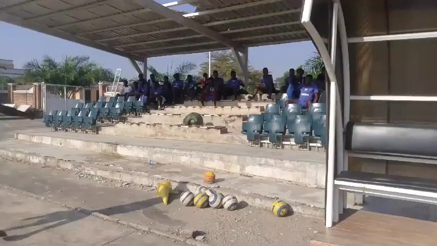 The video depicts a group of young men in blue shirts sitting in the stands of a small, outdoor stadium. The stands are under a metal awning, and there are rows of teal-colored seats. In front of the stands, there are several soccer balls and a yellow cone on the ground. The area appears to be a little run-down, with cracked concrete. In the background, there are palm trees and a building. A man in blue is walking around near the seated players.