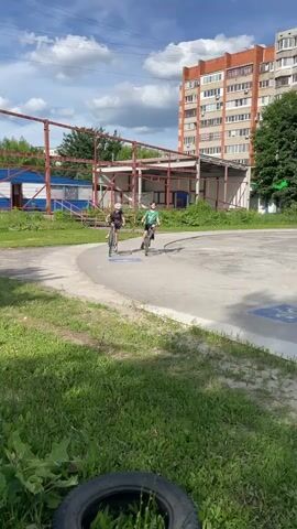 Two children are riding bicycles on a paved area. The child on the left is wearing a dark t-shirt, shorts, and a white helmet. The child on the right is wearing a green t-shirt, shorts, and a green helmet. They are riding side-by-side, and appear to be enjoying themselves. The background includes a large, multi-story apartment building, trees, and a sports facility with a red and blue structure. There is grass and a discarded tire in the foreground. The sky is blue with some fluffy white clouds, suggesting it is daytime and the weather is fair. The scene appears to be an outdoor recreational area in an urban or suburban setting, likely in Tula, Russia, given the architectural style of the apartment building. There are no visible signs or text.