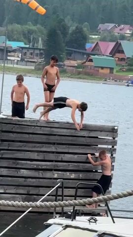 Two young males are on a wooden dock. One male, wearing black swim trunks, stands at the edge of the dock facing the water. Another male, also wearing black swim trunks, is attempting to climb the vertical side of the dock, his body inverted and facing the water. He appears to be struggling to maintain his grip. The dock is situated on a body of water, with houses and trees visible in the background. A boat is docked alongside the pier in the foreground.