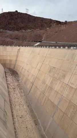 A drone camera pans over a large concrete dam, revealing a wide, dry channel running down the center of the dam face. The channel appears to be filled with debris, likely sediment and possibly vegetation. The dam walls are made of numerous concrete blocks, giving them a segmented appearance. The top of the dam is a flat walkway or road with railings. In the background, a rugged, brown mountain rises steeply, with a road winding up its side. Power lines and pylons are visible on the mountain's crest. Several vehicles are parked along the road on the mountain and on the dam's upper level. The sky is overcast with gray clouds. The camera's movement is smooth, and the perspective is an overhead, downward-looking view. There are no people clearly visible within the dam's channel, but there appear to be figures on the upper walkway of the dam and on the road in the background.