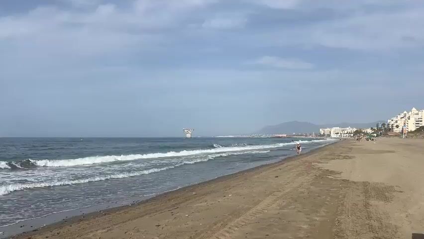 The video shows a wide view of a beach on a cloudy day. Gentle waves are breaking on the shore. The sand is mostly flat with some tire tracks visible. In the distance, there are buildings along the coastline, a few people walking on the beach, and what appears to be a large offshore structure. The ocean stretches out to the horizon under a pale sky.