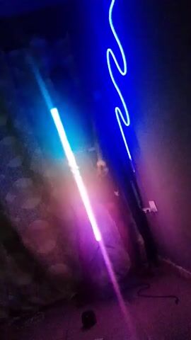 The video shows a person standing in a dark room next to a green neon light. The person is holding a light that is emitting multiple colors including blue, yellow, and orange. The room has textured wallpaper and is poorly lit, except for the light sources. The camera angle is slightly skewed, providing a tilted perspective of the scene.