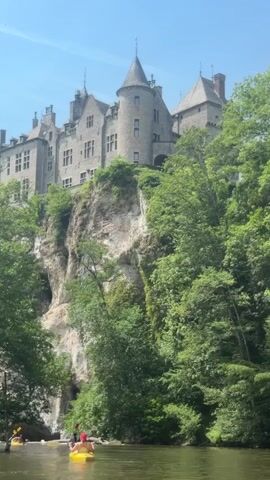 The video shows a wide shot of a river, with several kayaks on it.  The kayaks are bright yellow and appear to hold one or two people each, though their faces are indistinct.  Towering above the riverbank is a large, medieval-style castle made of grey stone.  The castle is built atop a steep cliff, with lush green trees surrounding its base and partially obscuring the view of the lower sections of the cliff.  The sky is clear and blue, indicating a bright, sunny day. The water is calm, and the overall scene suggests a peaceful day of kayaking in a picturesque location.