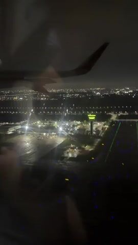 The video, filmed from the window of an airplane at night, shows an aerial view of an airport. An airplane wing is visible in the foreground, with lights reflecting off its surface. Below, the airport is illuminated by numerous lights. A control tower with red lights is prominent. Runways are visible with their characteristic lighting patterns. Buildings and parked vehicles can be seen on the tarmac. The distant cityscape is also visible, dotted with lights. The overall scene depicts an active airport operating at night.