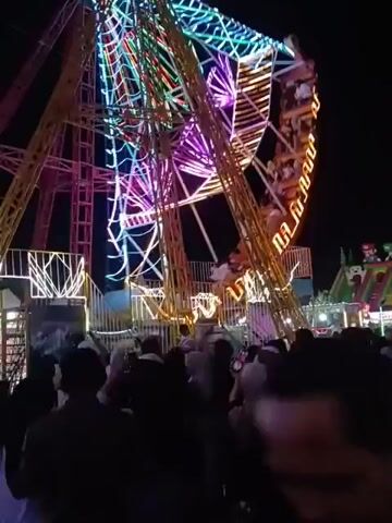 The video captures a nighttime scene at what appears to be a fair or carnival. The central focus is a large, illuminated amusement ride, possibly a Ferris wheel or a similar spinning attraction, adorned with numerous colorful lights in shades of pink, purple, green, and yellow. The ride is in motion, with several gondolas or seats visible, some occupied by people. Below the ride, a crowd of people is gathered, silhouetted against the bright lights of the attraction. They are observing the ride and the general activity of the fair. The environment is dark, indicating it is nighttime, and the lights from the ride are the primary source of illumination for the immediate area. Other carnival structures or attractions are faintly visible in the background. The overall atmosphere suggests a lively and festive event.