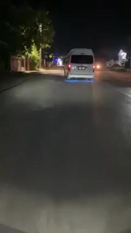 The video, taken at night, shows a street lit by overhead lights and car headlights. The camera is seemingly mounted on the front of a moving vehicle. The perspective is low, emphasizing the road surface.

At the beginning of the video, a white van with blue underglow is driving away in the distance. The road is paved and wet, reflecting the light. Trees and buildings line the sides of the street. As the video progresses, the vehicle with the camera moves forward, catching up with the van. The road is uneven and shows signs of wear. Street signs are visible on the side of the road.