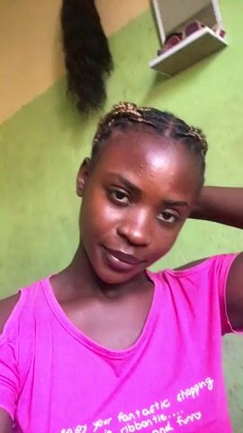 A young woman with dark skin and braided hair is looking directly at the camera. She is wearing a pink t-shirt with cut-out shoulders. The text on the shirt is difficult to read but appears to say something about "fantastic shopping". The background is a green wall with a shelf and some hair extensions visible in the upper corner. The woman is smiling and tilting her head slightly. The lighting appears to be natural daylight.