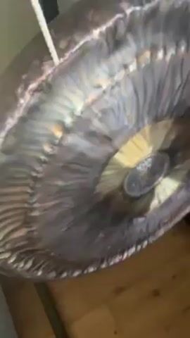 Here is a concise description of the video:

The video shows a close-up of a hanging gong. A white mallet strikes the surface of the gong in the video. The gong is metallic with swirling patterns and varying hues of silver, bronze, and copper. It's suspended by a white rope. The background includes a wooden floor and glimpses of a white wall.