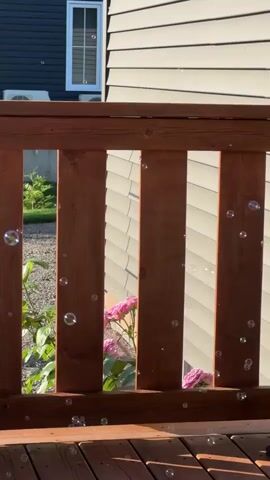A close-up, horizontal, eye-level video displays a wooden deck railing and gate in the foreground, with a dark blue house and a light tan house behind it. The wooden railing and gate are made of vertical planks of dark brown wood. Numerous small, clear, spherical bubbles are floating in the air and are visible through the gaps in the railing and on the deck surface. In the background, the dark blue house has two windows with white frames and blinds. Two outdoor air conditioning units are mounted on the side of the dark blue house. The light tan house has horizontal siding and a visible window with blinds. Pink roses are blooming behind the railing, and green foliage is visible. Sunlight casts shadows on the deck. The video pans slightly to the right over a duration of four seconds, revealing more of the deck and gate.