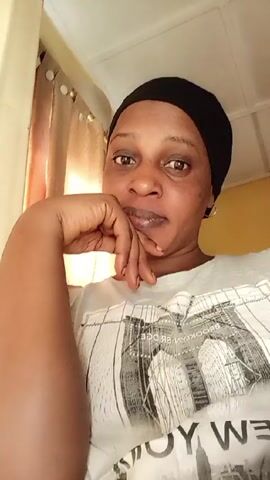 A woman in a black headscarf and light-colored t-shirt with a graphic of the Brooklyn Bridge is looking at the camera. She is resting her chin on her hand and smiling slightly. The background shows a window with sheer curtains and a glimpse of a yellow wall. The lighting is soft and natural.