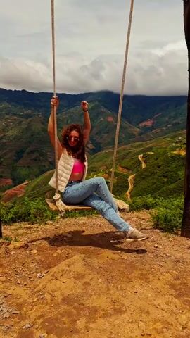 A young woman with curly red hair and sunglasses is swinging on a wooden swing suspended by ropes. She is wearing a pink crop top, a white quilted vest, and blue jeans. Her arms are raised in the air, and she is smiling. The swing is positioned on a grassy, rocky cliff overlooking a valley with terraced hillsides and mountains in the distance. The sky is cloudy.
